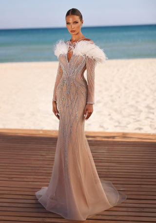 Woman in a long, beaded gown with feather accents, standing near a beach.